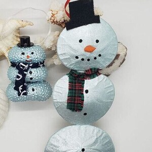 Op de afbeelding: Twee blauwe zanddollar-sneeuwman ornamenten. De grotere sneeuwman heeft een zwarte hoed, een rood-groene geruite sjaal en een wortelneus. De kleinere sneeuwman heeft een zwarte hoed, een blauw-witte gestreepte sjaal en een zwarte knoopneus.