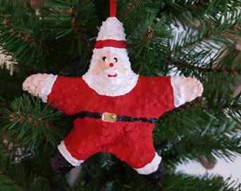 May include: A red and white Santa Claus shaped Christmas ornament. The ornament is made of paper mache and has a white beard and a black belt.