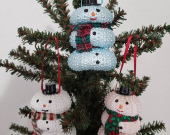 May include: Three snowman ornaments made from sea urchins. The snowmen are decorated with black hats, red and white scarves, and black eyes and buttons. The snowmen are hanging on a green Christmas tree.