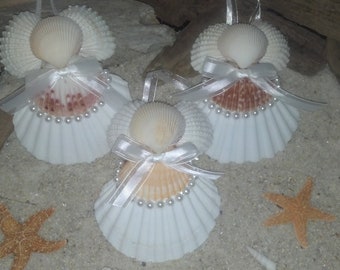 May include: Three seashell angel ornaments with white shell wings, pearl accents, and a small white bow. The angels are on a bed of sand with starfish and driftwood.