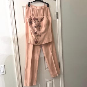 May include: A peach-colored two-piece set consisting of a sleeveless camisole top and matching trousers. The top features thin spaghetti straps and a decorative floral appliqué design with fabric petals cascading down the front left side.
