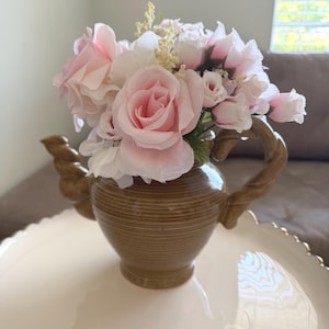 May include: A brown ceramic teapot with a twisted handle and spout, filled with a bouquet of artificial light pink roses and small white filler flowers. The teapot has a rounded body with horizontal ridges and a smooth, glossy finish.
