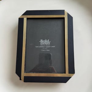 May include: A rectangular photo frame featuring a matte black border accented with polished gold-tone brass trim. The center insert displays the text Solid Brass Photo Frame, hand polished & lacquer coated, 5 inches by 7 inches, 13cm by 18cm.