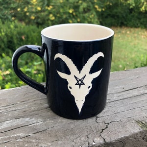 May include: A black ceramic coffee mug with a white goat head and pentagram design.