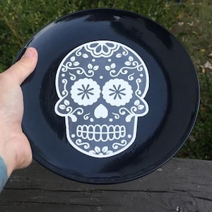 Könnte beinhalten: Ein schwarzer Keramikteller mit einem weißen Zuckerschädel-Design. Der Schädel hat florale und wirbelnde Details, mit zwei blütenförmigen Augen und einer herzförmigen Nase. Der Teller wird von einer Hand gehalten.