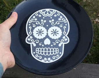 May include: A black ceramic plate featuring a white sugar skull design. The skull has floral and swirl details, with two flower-shaped eyes and a heart-shaped nose. The plate is held by a hand.