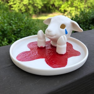 Peut inclure: Porte-bague en céramique blanche en forme d'agneau avec une larme bleue sur le visage. L'agneau est couché dans une flaque de peinture rouge sur un plat en céramique blanche.