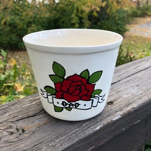 Könnte beinhalten: Weißer Keramiktopf mit einer roten Rose und grünen Blättern, ein weißes Band mit dem Wort "DAD" in schwarzen Buchstaben.