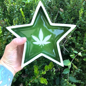 Könnte beinhalten: Eine sternförmige Keramikschale mit glänzend grüner Innenseite und weißem Rand. In der Mitte befindet sich ein weißes Cannabisblatt-Design. Die Schale wird vor einem Hintergrund aus grünem Laub gehalten.