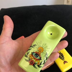 Könnte beinhalten: Eine hellgrüne Keramikpfeife mit einem dekorativen Design mit einem Auge in einem Dreieck, einer Sonne und floralen Elementen. Die Pfeife ist rechteckig mit einer Schale und einem Loch. Eine gelbe Pfeife mit Fischmuster ist ebenfalls sichtbar.