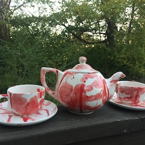 Op de afbeelding: Een witte theepot en twee theekopjes met een rood splatter-ontwerp. De theepot heeft een rond lichaam en een deksel. De theekopjes hebben een handvat en een schoteltje.
