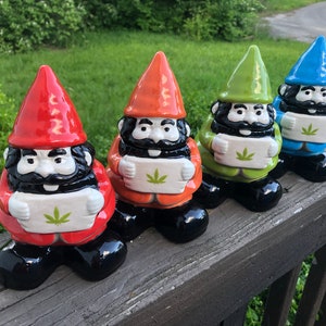 Puede incluir: Cuatro gnomos de cerámica sosteniendo un cartel blanco con una hoja de marihuana verde. Los gnomos llevan sombreros rojos, naranja, verde y azul. Los gnomos están de pie sobre una superficie de madera.