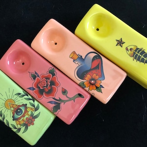 May include: Four ceramic pipes in green, pink, and yellow, each featuring a tattoo-style design. Designs include a rose, a bottle with a heart, a fish skeleton, and an eye within a triangle. The pipes are rectangular with a bowl.