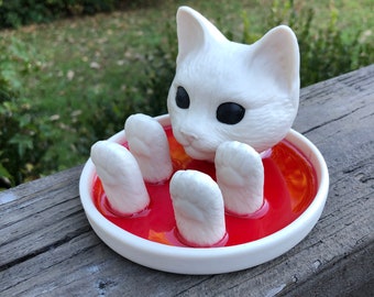 May include: White ceramic cat figurine with black eyes, sitting in a dish filled with red liquid. The cat's paws are submerged in the liquid.