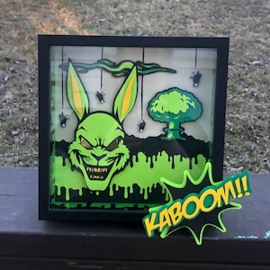 May include: A framed, layered art piece featuring a menacing green rabbit with yellow accents, a mushroom cloud, and the word "KABOOM!!" in a comic book style. The artwork has a black frame and a green and black color scheme.