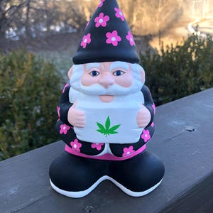 May include: A black and pink garden gnome with a white beard and a pink flower hat. The gnome is holding a white sign with a green marijuana leaf.