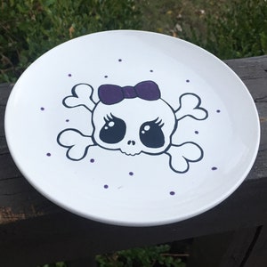 Könnte beinhalten: Ein weißer Keramikteller mit einem schwarzen und weißen Totenkopfdesign. Der Totenkopf hat violette Augen und eine violette Schleife. Der Teller ist mit kleinen violetten Punkten verziert.