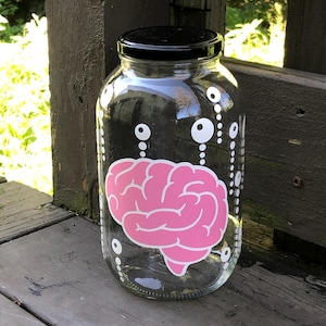 以下が含まれることがあります： 黒い蓋が付いた透明なガラス瓶で、前面にピンク色の脳のグラフィックが描かれています。瓶には白い円形のアクセントと、上部から下部に向かって伸びる垂直の点線が装飾されており、独特の視覚的なパターンを作り出しています。