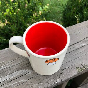 May include: White ceramic mug with a red interior and a mushroom design. The mug has a handle and is sitting on a wooden surface.
