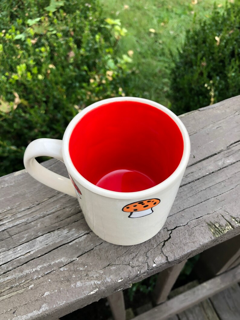 May include: White ceramic mug with a red interior and a mushroom design. The mug has a handle and is sitting on a wooden surface.