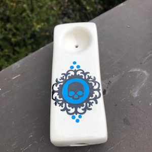 May include: A white ceramic pipe with a blue and black skull design. The pipe has a rectangular shape with a bowl at the top and a hole at the bottom. The skull is inside a blue circle with a decorative border.