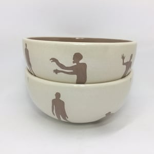 Puede incluir: Dos cuencos de cerámica blanca con siluetas marrones de personas. Los cuencos están apilados uno encima del otro.