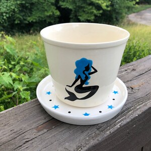 Op de afbeelding: Een witte keramische plantenpot met een blauw zeemeermin silhouet en blauwe sterren op de schotel. De pot staat op een houten oppervlak.
