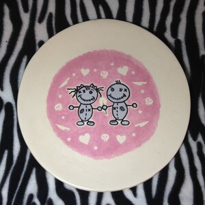 May include: A white ceramic plate with a pink background featuring two cartoon figures holding hands. The figures are gray with black eyes and mouths. The background is decorated with hearts, stars, and skulls.