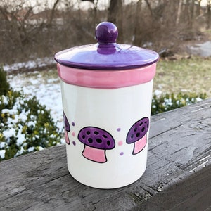 以下が含まれることがあります： 紫色の蓋とピンクの縁取りが付いた白い陶器の壺。壺には、ピンクの茎と白い水玉模様の紫色のキノコのデザインが施されています。