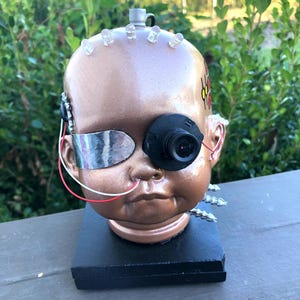 May include: A modified doll head with a bronze finish, featuring a camera eye, LED lights, and metallic accents. The doll head is mounted on a black base, creating a unique, artistic piece.