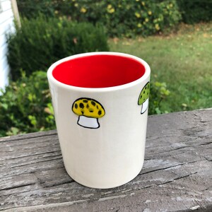May include: White ceramic mug with a red interior and two hand-painted mushroom designs. One mushroom is yellow with black spots and the other is green with white spots.