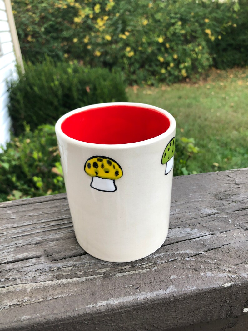 May include: White ceramic mug with a red interior and two hand-painted mushroom designs. One mushroom is yellow with black spots and the other is green with white spots.