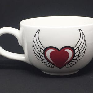 Könnte beinhalten: Weiße Keramik-Kaffeetasse mit einem großen roten Herz und schwarzen Flügeln. Die Tasse hat eine abgerundete Form und einen gebogenen Henkel. Das Herz- und Flügeldesign ist mittig auf der Vorderseite der Tasse angebracht.
