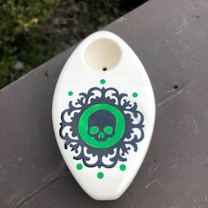 May include: Oval-shaped, off-white ceramic incense holder with a black and green skull design. The design features a black skull in a green circle, surrounded by a black floral pattern and green dots. The top has a small hole for incense.