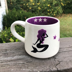 May include: A white ceramic mug with a purple interior and a black silhouette of a mermaid with purple hair. The mug has five white stars on the inside rim and two white stars on the outside.