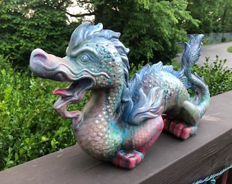 May include: A ceramic dragon sculpture in shades of blue, green, and red. The dragon has an open mouth, detailed scales, and decorative accents. It is positioned on a wooden surface with a natural background of greenery.