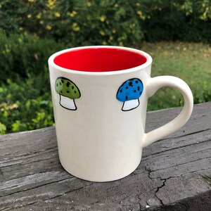 May include: White ceramic mug with a red interior and two painted mushroom designs. One mushroom is green with black spots and the other is blue with black spots.