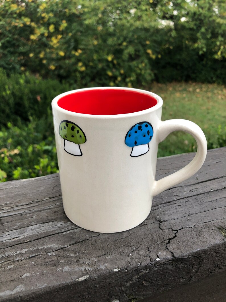 May include: White ceramic mug with a red interior and two painted mushroom designs. One mushroom is green with black spots and the other is blue with black spots.