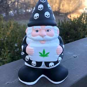 以下が含まれることがあります： 骸骨柄の帽子をかぶった黒と白の庭のノームが、緑色のマリファナの葉の描かれた看板を持っている。