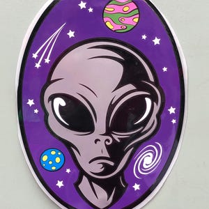 May include: An oval sticker featuring a gray alien head centered against a deep purple space background. The design includes a green and pink striped planet, a blue planet with yellow dots, a white spiral galaxy, and several white stars.