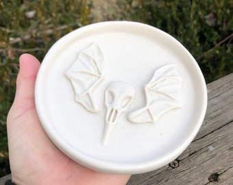 May include: White ceramic dish with a raised design of a skull with wings. The dish is approximately 4 inches in diameter.