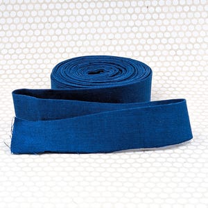 May include: A roll of deep blue fabric ribbon with a visible woven texture. The ribbon is coiled into a tight cylinder with one end extending outward, displaying a consistent dark blue color and a flat, straight-edged design throughout.