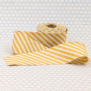 May include: A roll of fabric ribbon featuring a repeating diagonal stripe pattern in mustard yellow and off-white. The ribbon is wound into a tight spool, with a length of the striped textile extending outward to display the consistent color scheme.
