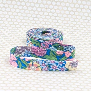 May include: A roll of fabric ribbon featuring a vibrant floral pattern with pink, blue, yellow, and green flowers on a bright blue background. The ribbon is coiled into a circular shape with a loose end extending forward.
