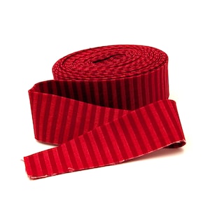 May include: A spool of red ribbon featuring a repeating vertical striped pattern. The ribbon is tightly wound into a cylindrical shape with one end extending outward, showcasing the consistent texture and deep red color of the fabric material.