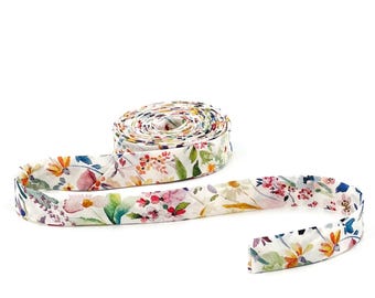 May include: A roll of white fabric bias tape featuring a colorful watercolor floral pattern with shades of pink, orange, yellow, blue, and green. The tape is coiled into a circular spool with one end extending outward to display the print.