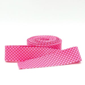 May include: A roll of bright pink ribbon featuring a repeating red diamond grid pattern. The ribbon is coiled into a circular spool with one end extending outward, displaying the consistent geometric lattice design across the entire surface.