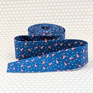 May include: A roll of blue fabric with a pattern of small American flags and white stars. The fabric is unrolled in the foreground, revealing the pattern. The background is a light beige dotted pattern.
