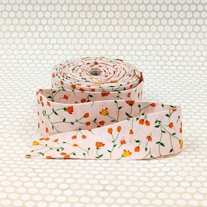May include: A roll of light pink fabric ribbon featuring a repeating pattern of small orange and yellow flowers with thin green stems and leaves. The ribbon is coiled into a circular shape with one end extending outward across the surface.