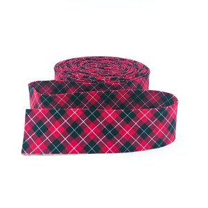 May include: A roll of fabric ribbon featuring a diagonal plaid pattern in shades of red, black, and white. The ribbon is coiled into a circular shape with one end extending outward, displaying the consistent geometric tartan design throughout.